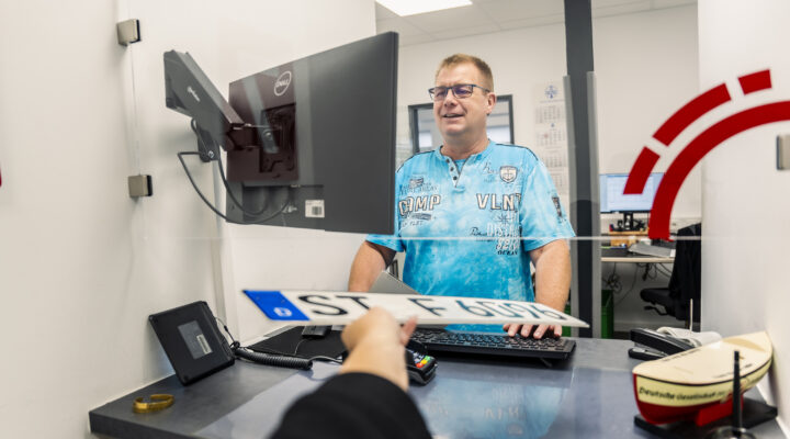 Mitarbeiter hinter einer Glasscheibe am Serviceschalter, trägt ein blaues T-Shirt und spricht mit einer Person auf der anderen Seite. Vor ihm stehen Monitor, Tastatur und Kartenlesegerät; auf dem Tresen liegt ein Ausweis oder Dokument.