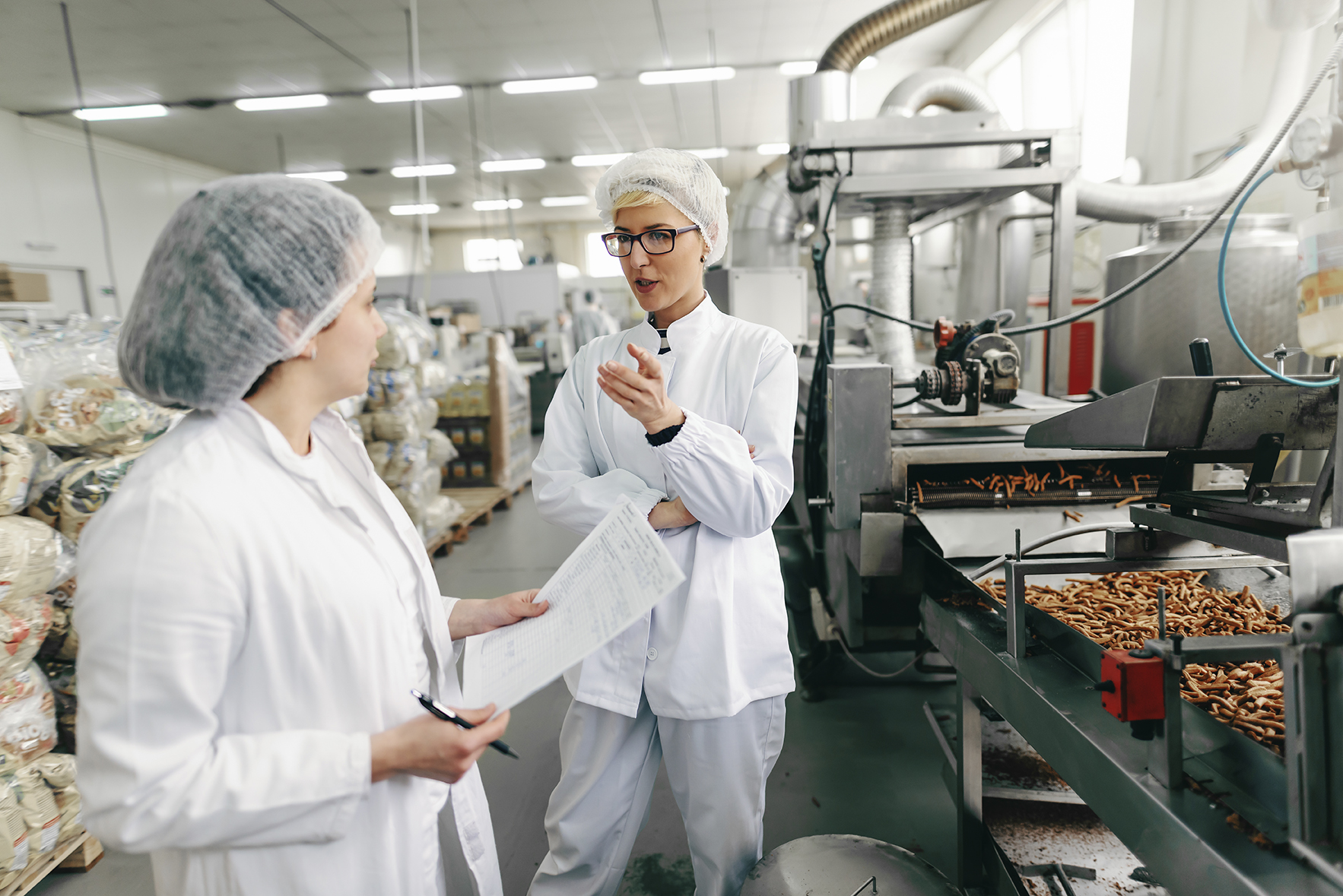 Lebensmittelkontrolleurin und Fabrikmitarbeiterin in weißen Uniformen und mit sterilen Hauben auf den Köpfen unterhalten sich über die Hygienequalität, während sie in einer Lebensmittelfabrik stehen.