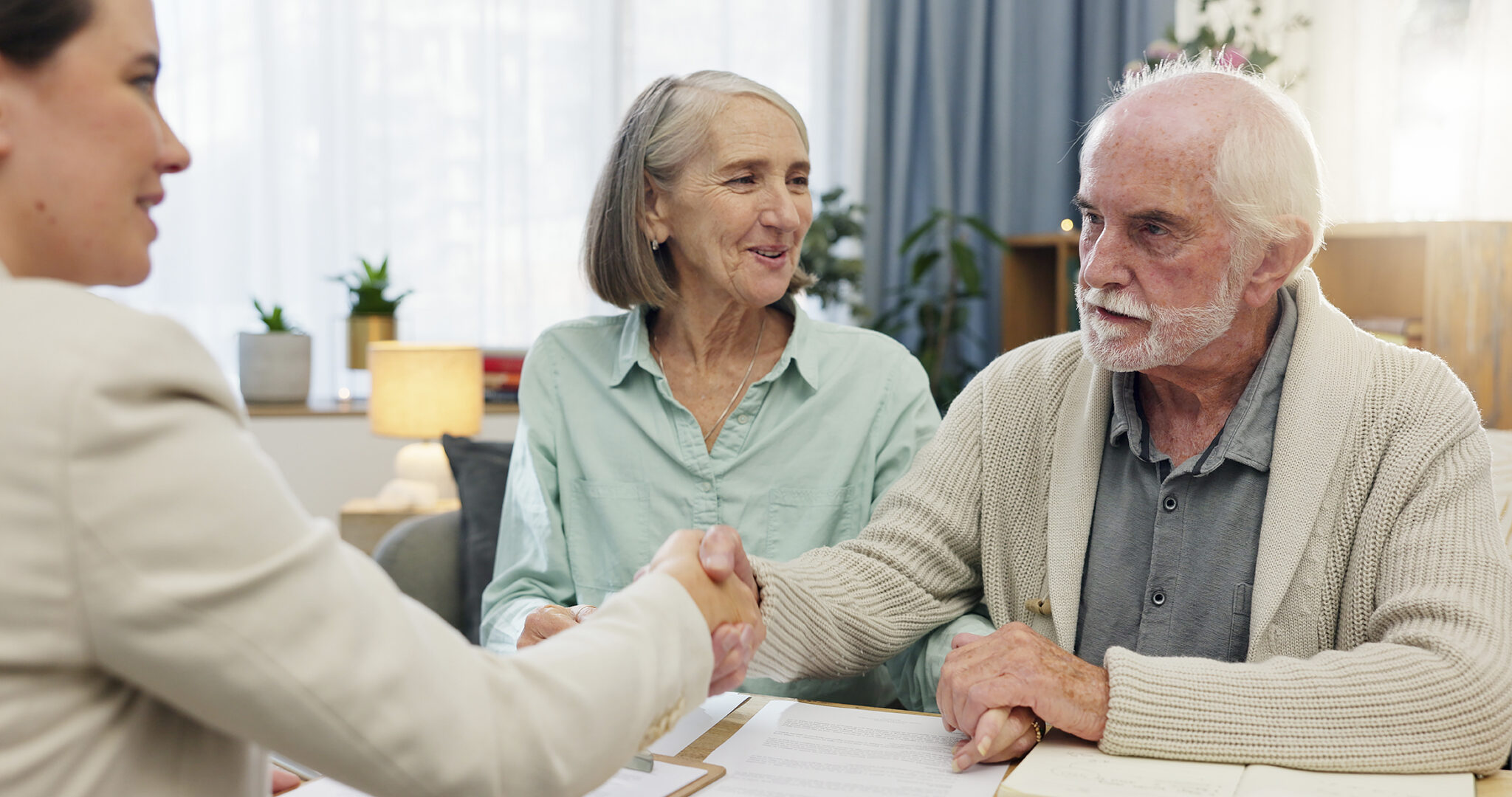 Beratungsgespräch zu Hause mit älterem Ehepaar und Beraterin – Händeschütteln als Zeichen einer Einigung zu Ruhestandsplanung, Testament oder rechtlicher Vorsorge.