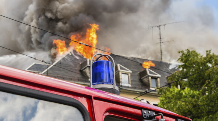 Dach eines mehrstöckigen Gebäudes in Flammen, aus dem dichter schwarzer Rauch aufsteigt, im Vordergrund das Blaulicht eines roten Feuerwehrfahrzeugs.
