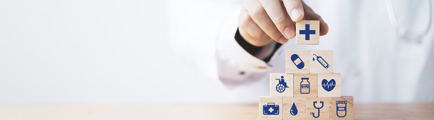 Person im weißen Arztkittel mit Stethoskop stapelt Holzklötze mit medizinischen Symbolen wie Kreuz, Spritze, Herz, Rollstuhl und Medikamenten.
