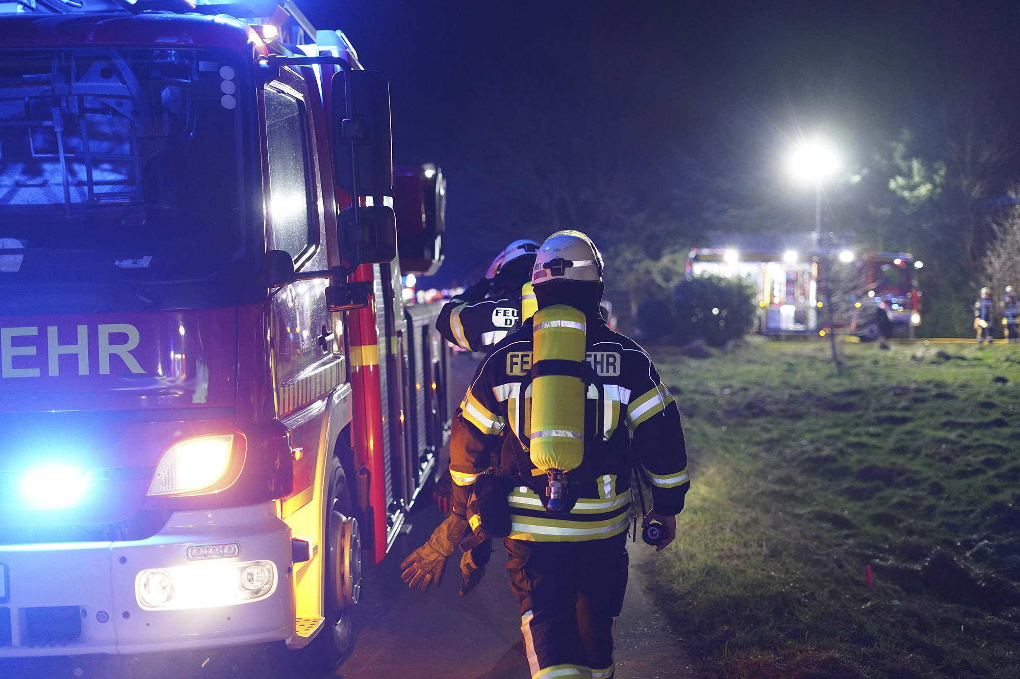 Feuerwehrleute in voller Schutzausrüstung mit Atemgeräten gehen nachts an einem Einsatzfahrzeug vorbei, im Hintergrund weitere beleuchtete Feuerwehrfahrzeuge und Einsatzkräfte auf einer Wiese.