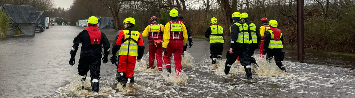 Einsatzkräfte in Schutzanzügen und Warnwesten waten durch eine überflutete Straße. Im Hintergrund sind Bäume und Einsatzfahrzeuge zu sehen, die auf einen Hochwassereinsatz hinweisen.