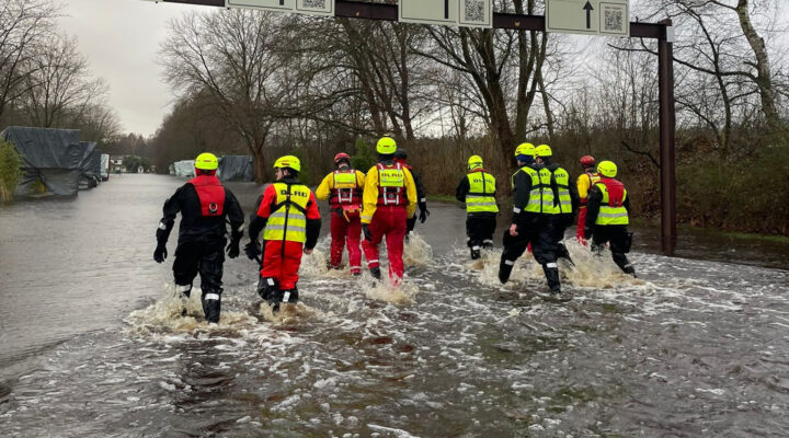 Einsatzkräfte in Schutzanzügen und Warnwesten waten durch eine überflutete Straße. Im Hintergrund sind Bäume und Einsatzfahrzeuge zu sehen, die auf einen Hochwassereinsatz hinweisen.