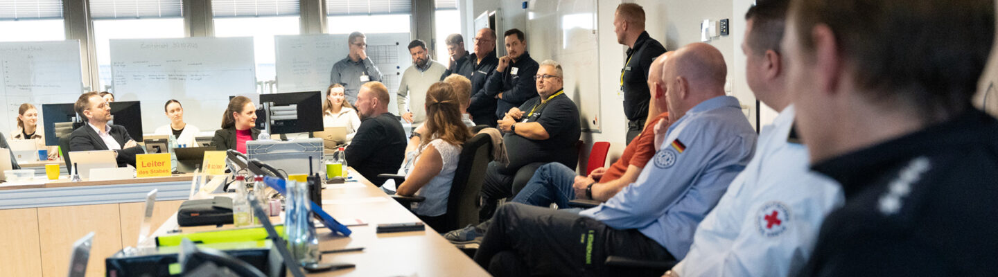 Katastrophenschutzübung: Mehrere Einsatzkräfte und Mitarbeitende verschiedener Organisationen sitzen und stehen in einem Besprechungsraum. Auf den Tischen liegen Unterlagen, Telefone und Computer. Im Hintergrund sind Whiteboards mit Notizen zu sehen.
