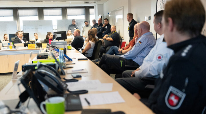 Katastrophenschutzübung: Mehrere Einsatzkräfte und Mitarbeitende verschiedener Organisationen sitzen und stehen in einem Besprechungsraum. Auf den Tischen liegen Unterlagen, Telefone und Computer. Im Hintergrund sind Whiteboards mit Notizen zu sehen.