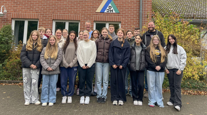 Eine Gruppe von Jugendlichen und zwei erwachsenen Begleitpersonen steht in mehreren Reihen vor einem Backsteingebäude. Alle blicken in die Kamera und tragen herbstliche Kleidung. Über dem Eingang ist ein Schild mit dem DJH-Logo zu sehen, im Hintergrund befinden sich Büsche und Bäume.