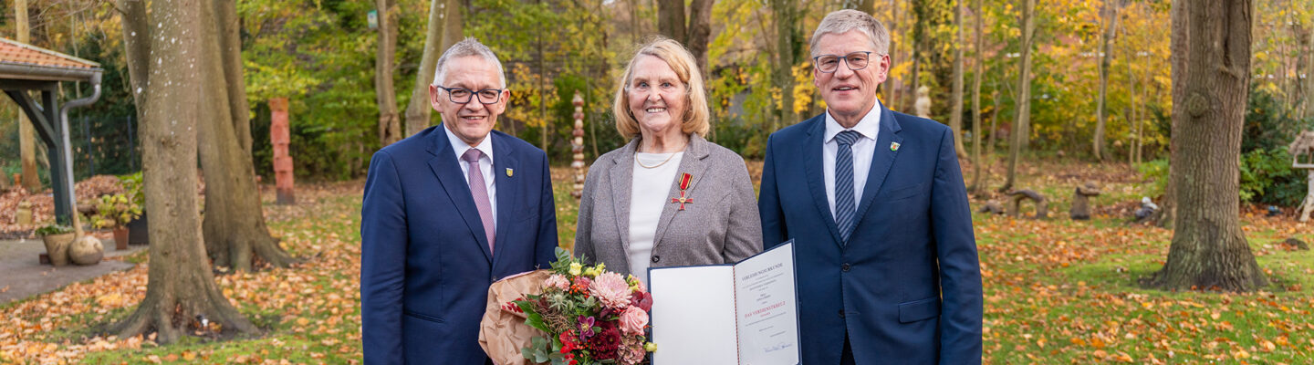 Landrat Olaf Meinen, geehrte Anna Uphoff und Bürgermeister Horst Feddermann stehen im Freien auf einem herbstlich bedeckten Weg. Anna Uphoff in der Mitte hält einen Blumenstrauß und eine geöffnete Urkunde, während Olaf Meinen und Horst Feddermann links und rechts von ihr stehen und lächeln. Im Hintergrund sind Bäume mit herabgefallenen Blättern zu sehen.