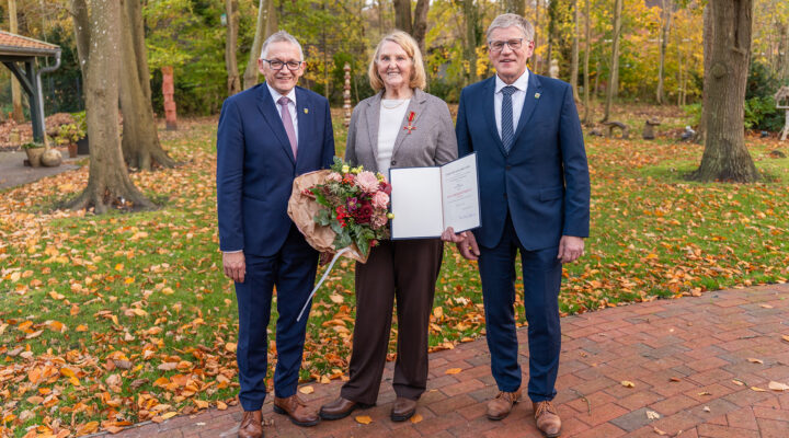 Landrat Olaf Meinen, geehrte Anna Uphoff und Bürgermeister Horst Feddermann stehen im Freien auf einem herbstlich bedeckten Weg. Anna Uphoff in der Mitte hält einen Blumenstrauß und eine geöffnete Urkunde, während Olaf Meinen und Horst Feddermann links und rechts von ihr stehen und lächeln. Im Hintergrund sind Bäume mit herabgefallenen Blättern zu sehen.