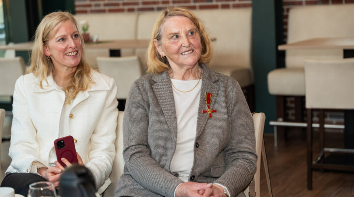 Anna Uphoff und ihre Tochter sitzen in einem modern eingerichteten Raum nebeneinander und lächeln. Anna Uphoff trägt ein Jackett und das Bundesverdienstkreuzes an ihrer Brust; die Tochter links hält ein Smartphone in der Hand. Im Hintergrund stehen helle Stühle und Tische.