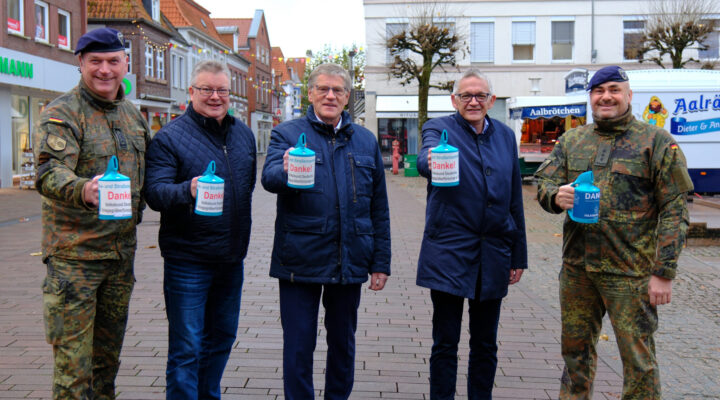 Fünf Männer stehen in einer Einkaufsstraße und halten jeweils eine Sammeldose mit der Aufschrift ‚Danke!‘ in der Hand. Zwei der Männer tragen Bundeswehruniformen, die drei anderen dunkle Jacken. Im Hintergrund sind Geschäfte und ein Brötchenwagen zu sehen.