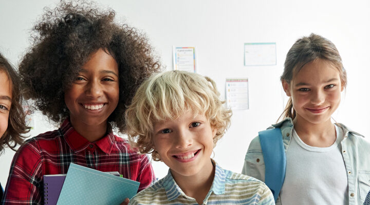 Fünf fröhlich lächelnde Schulkinder unterschiedlichen Aussehens stehen nebeneinander in einem hellen Klassenraum. Zwei von ihnen tragen Rucksäcke, eines hält Hefte in den Armen. Im Hintergrund hängen bunte Lernplakate an der Wand.