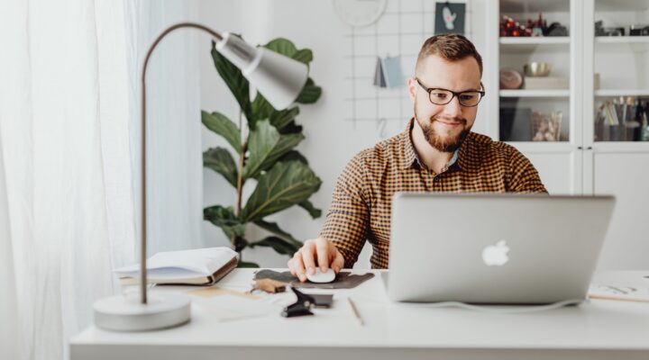 Ein Mann sitzt an einem hellen Schreibtisch in einem modernen Arbeitszimmer und arbeitet an einem Laptop. Neben ihm stehen eine Tischlampe, Notizunterlagen und ein Smartphone; im Hintergrund sind Pflanzen und ein Regal mit Büchern zu sehen.