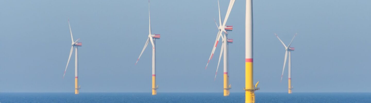 Mehrere Offshore-Windkraftanlagen stehen im Meer; ihre weißen Rotorblätter drehen sich vor einem klaren blauen Himmel, die Fundamente ragen gelb aus dem Wasser.