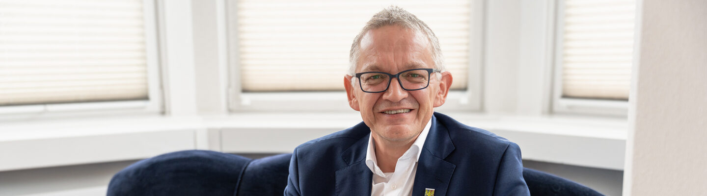 Landrat Olaf Meinen sitzt lächelnd auf einem roten Sofa in einem hellen Raum. Er trägt einen dunkelblauen Anzug mit weißem Hemd und Brille; hinter ihm sind Fenster mit Jalousien zu sehen.