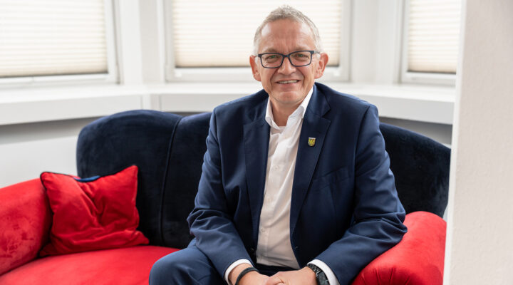 Landrat Olaf Meinen sitzt lächelnd auf einem roten Sofa in einem hellen Raum. Er trägt einen dunkelblauen Anzug mit weißem Hemd und Brille; hinter ihm sind Fenster mit Jalousien zu sehen.