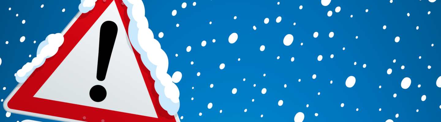 Verkehrszeichen mit rotem Warn-Dreieck und Ausrufezeichen, darunter ein Zusatzschild mit Schneeflocken-Symbol. Das Schild ist mit Schnee bedeckt, im Hintergrund fällt dichter Schnee vor blauem Himmel – Hinweis auf winterliche Glätte.