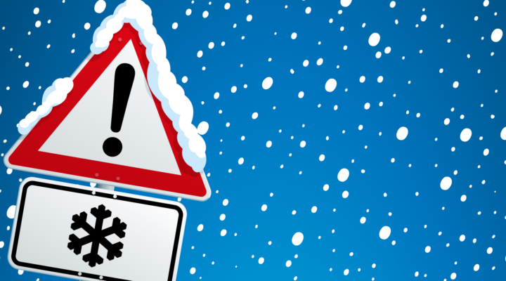 Verkehrszeichen mit rotem Warn-Dreieck und Ausrufezeichen, darunter ein Zusatzschild mit Schneeflocken-Symbol. Das Schild ist mit Schnee bedeckt, im Hintergrund fällt dichter Schnee vor blauem Himmel – Hinweis auf winterliche Glätte.
