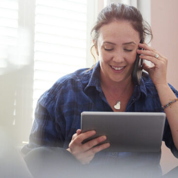 Eine Frau sitzt in einem hellen Raum, telefoniert mit einem Smartphone und hält gleichzeitig ein Tablet in der Hand. Sie lächelt, während Tageslicht durch ein Fenster mit Lamellen fällt; der Vordergrund ist unscharf.