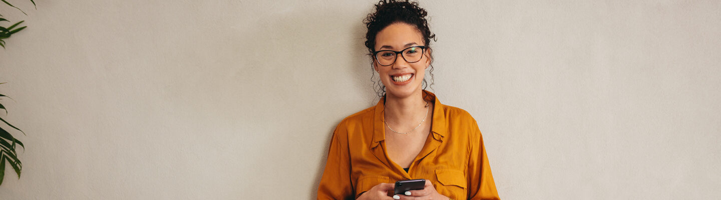 „Eine Frau steht lächelnd vor einer hellen Wand und hält ein Smartphone mit beiden Händen. Sie trägt eine orangefarbene Bluse, eine Jeans und eine Brille; links ist eine Zimmerpflanze angeschnitten zu sehen.