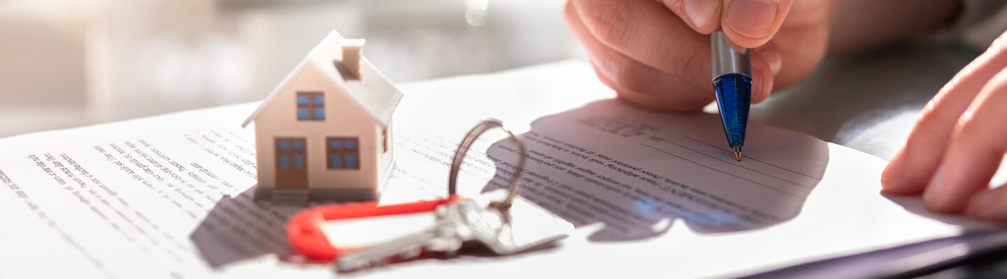 Eine Hand unterschreibt mit einem Stift ein Dokument. Auf dem Papier liegen ein kleines Modellhaus und ein Schlüsselbund – Symbol für einen Hauskauf, Mietvertrag oder eine Immobilienvereinbarung.