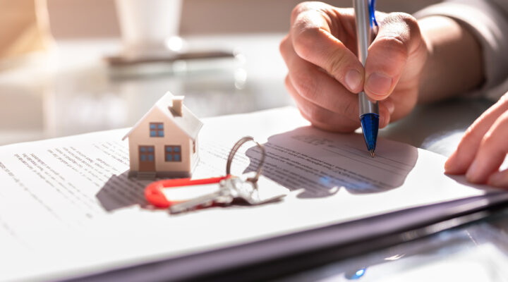 Eine Hand unterschreibt mit einem Stift ein Dokument. Auf dem Papier liegen ein kleines Modellhaus und ein Schlüsselbund – Symbol für einen Hauskauf, Mietvertrag oder eine Immobilienvereinbarung.