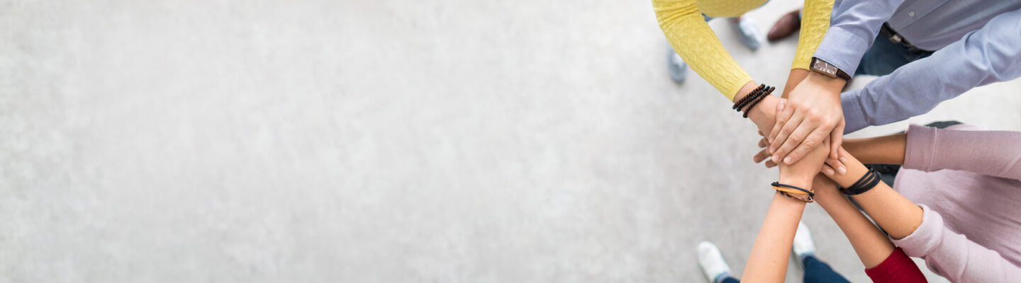 Mehrere Personen legen ihre Hände übereinander in der Mitte eines Kreises. Die Aufnahme ist von oben gemacht und symbolisiert Zusammenhalt, Teamarbeit und gegenseitige Unterstützung.