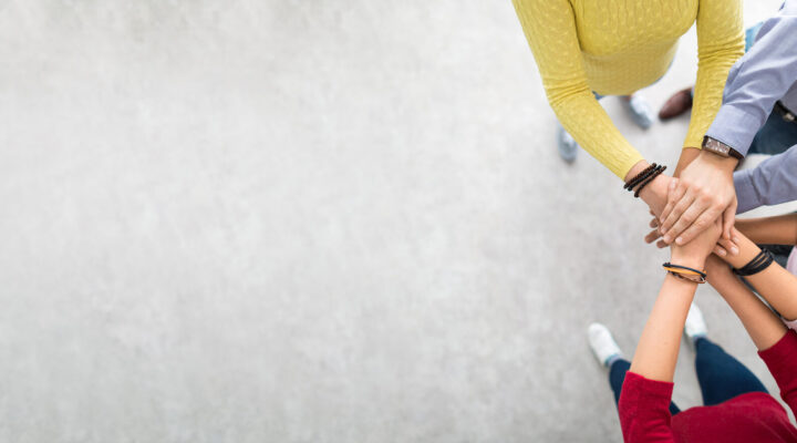 Mehrere Personen legen ihre Hände übereinander in der Mitte eines Kreises. Die Aufnahme ist von oben gemacht und symbolisiert Zusammenhalt, Teamarbeit und gegenseitige Unterstützung.