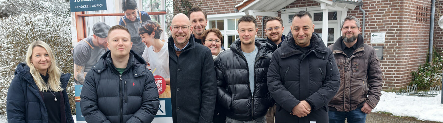 Eine Gruppe von Erwachsenen steht im Freien vor einem Backsteingebäude. Neben ihnen ist ein Roll-up-Banner des Jobcenters Landkreis Aurich aufgestellt. Die Personen tragen winterliche Kleidung; im Hintergrund sind Schnee, Bäume und der Gebäudeeingang zu sehen.