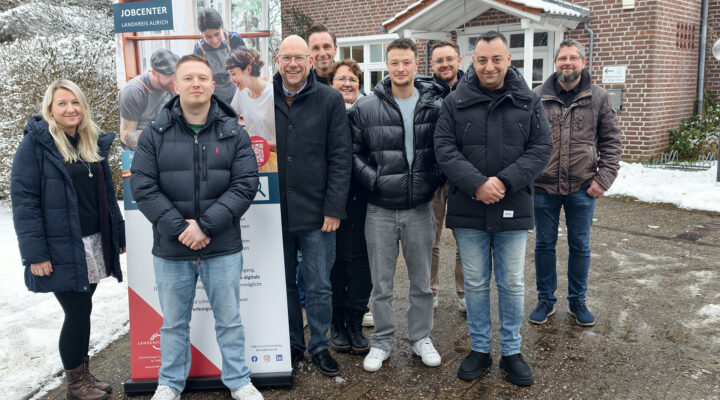 Eine Gruppe von Erwachsenen steht im Freien vor einem Backsteingebäude. Neben ihnen ist ein Roll-up-Banner des Jobcenters Landkreis Aurich aufgestellt. Die Personen tragen winterliche Kleidung; im Hintergrund sind Schnee, Bäume und der Gebäudeeingang zu sehen.