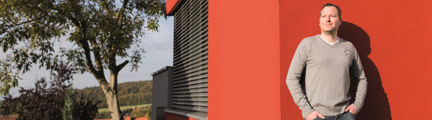 Eine Person steht an der Außenwand eines modernen, rot gestrichenen Gebäudes und blickt seitlich in die Sonne. An der Wand ist ein grünes Schild mit der Zahl 27 angebracht. Im Hintergrund sind Bäume und eine ländliche Landschaft zu sehen.