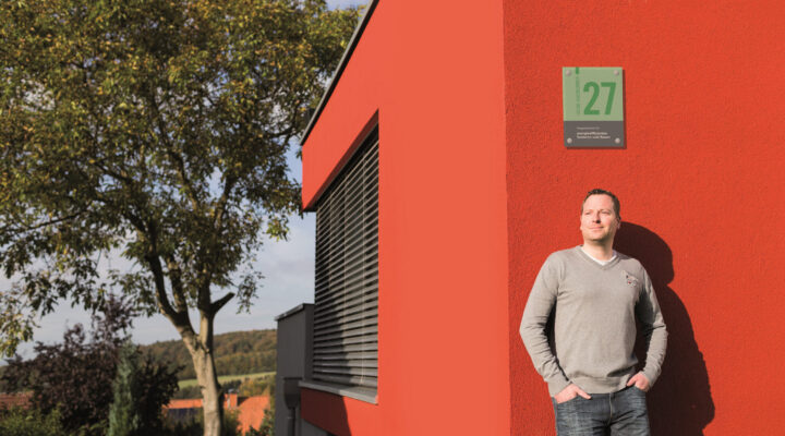 Eine Person steht an der Außenwand eines modernen, rot gestrichenen Gebäudes und blickt seitlich in die Sonne. An der Wand ist ein grünes Schild mit der Zahl 27 angebracht. Im Hintergrund sind Bäume und eine ländliche Landschaft zu sehen.